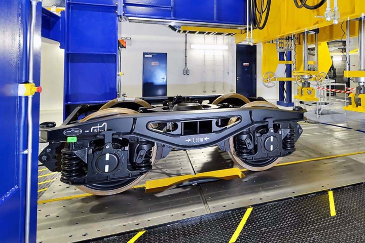 Railway bogie mounted on testing rig inside engineering laboratory, with blue and yellow machinery and industrial equipment visible.